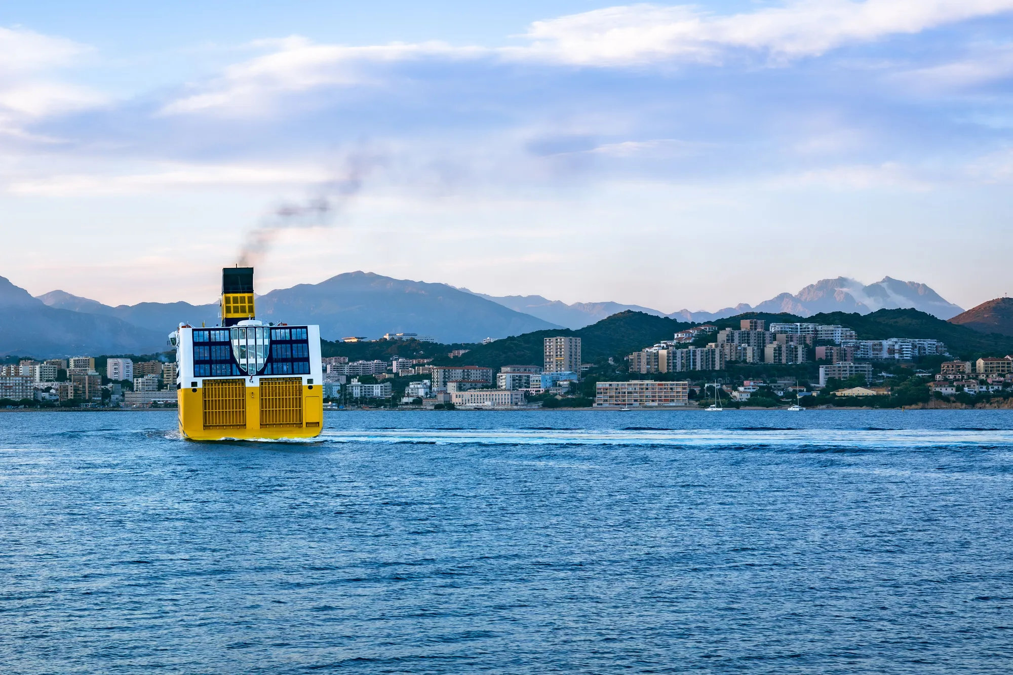 Un ferry jaune arrive à Ajaccio, la ville et les montagnes au fond